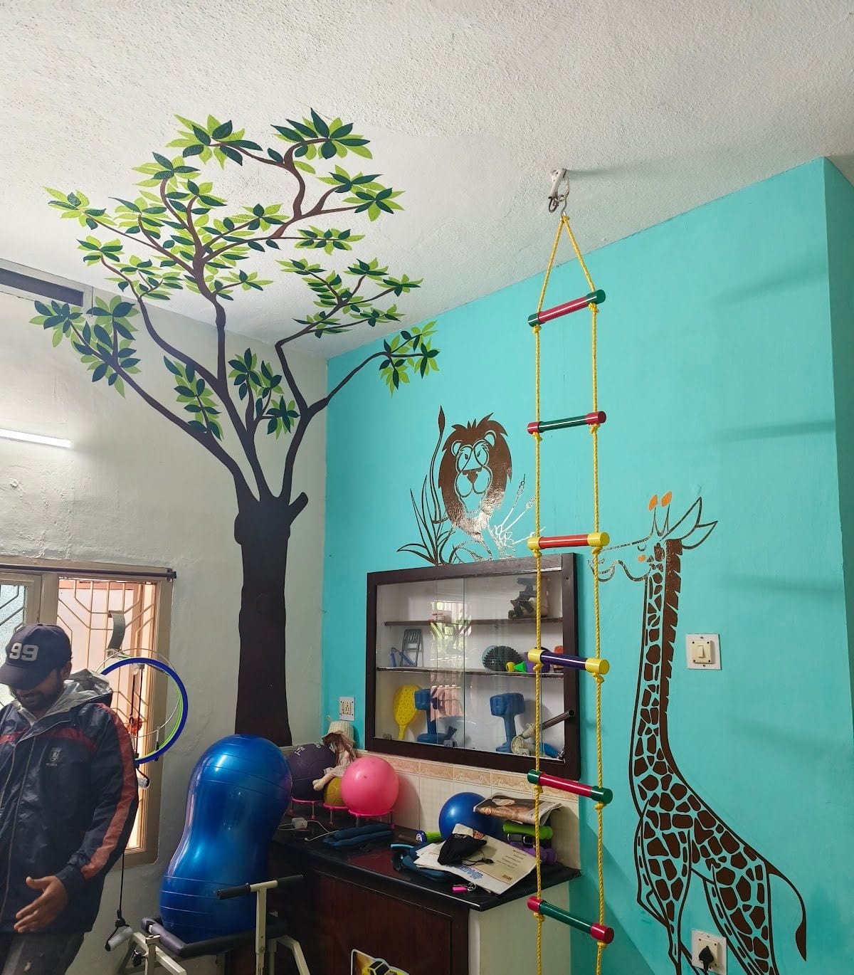 Play-based therapy room with tree and animal wall murals, a climbing rope ladder, gym balls and an equipment cabinet